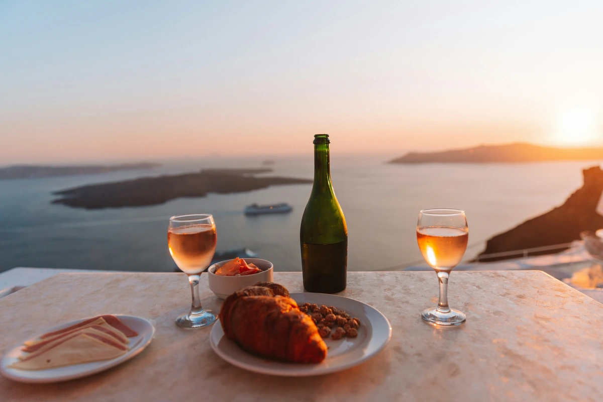 Duas taças de vinho, uma garrafa e pratos com queijos e pães sobre uma mesa diante do mar ao pôr do sol.