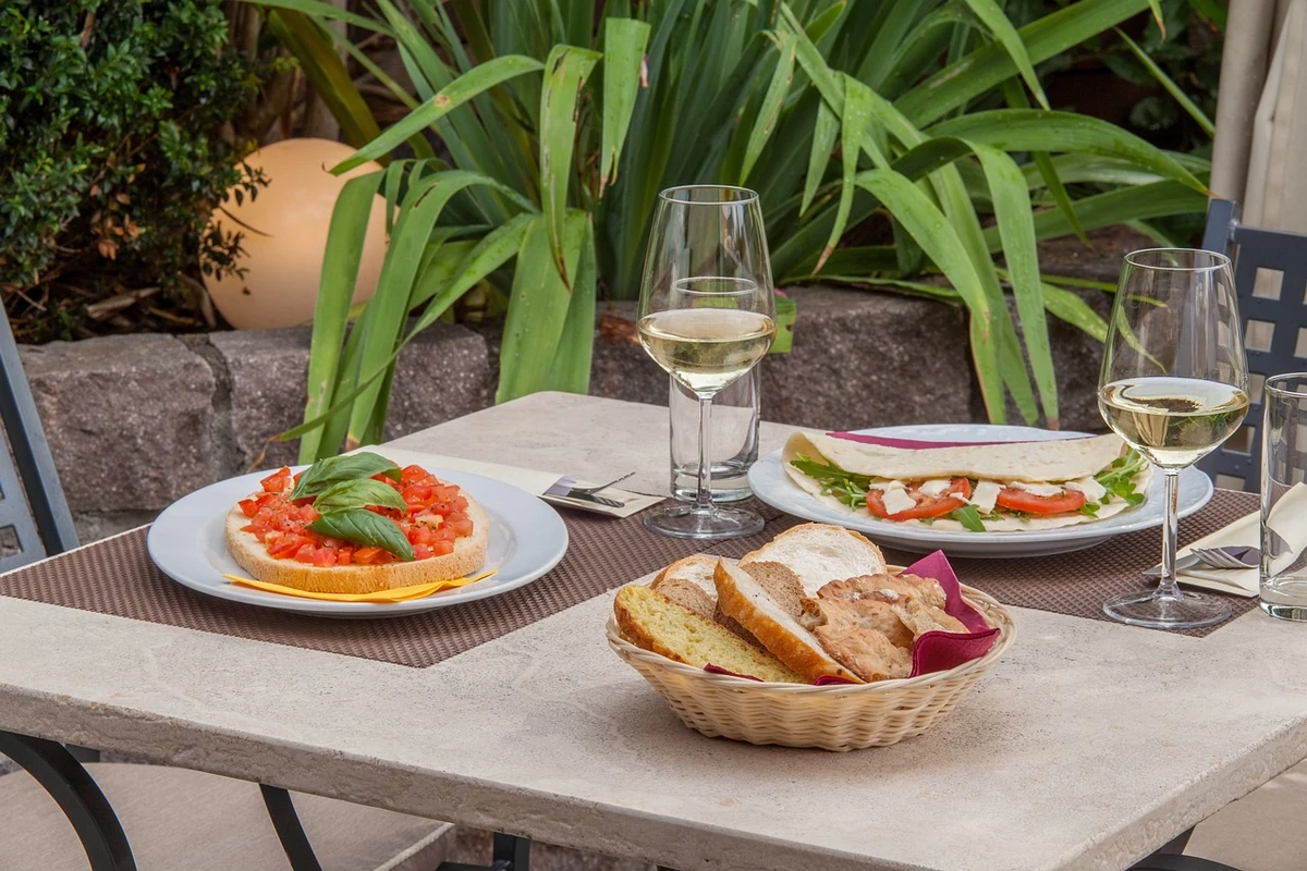 Mesa ao ar livre com bruschetta de tomate e manjericão, sanduíche, cesta de pães e duas taças de vinho claro.