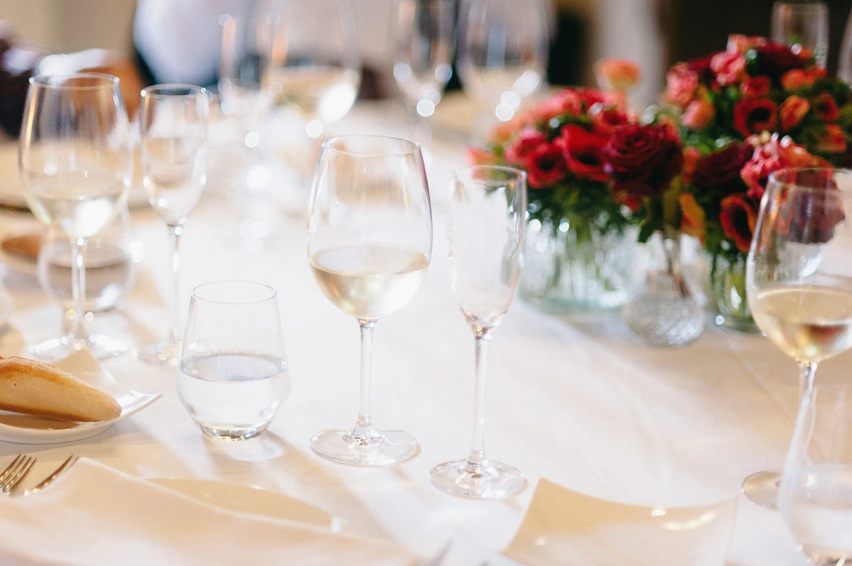 Taça de vinho branco no centro de uma mesa com toalha branca e arranjo de flores.