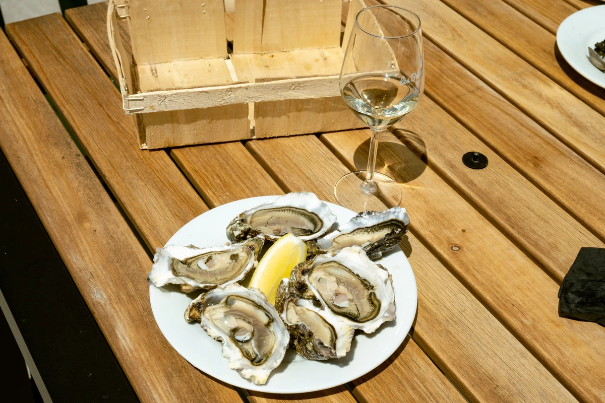 Prato de ostras com limão ao lado de uma taça de vinho branco sobre mesa de madeira.