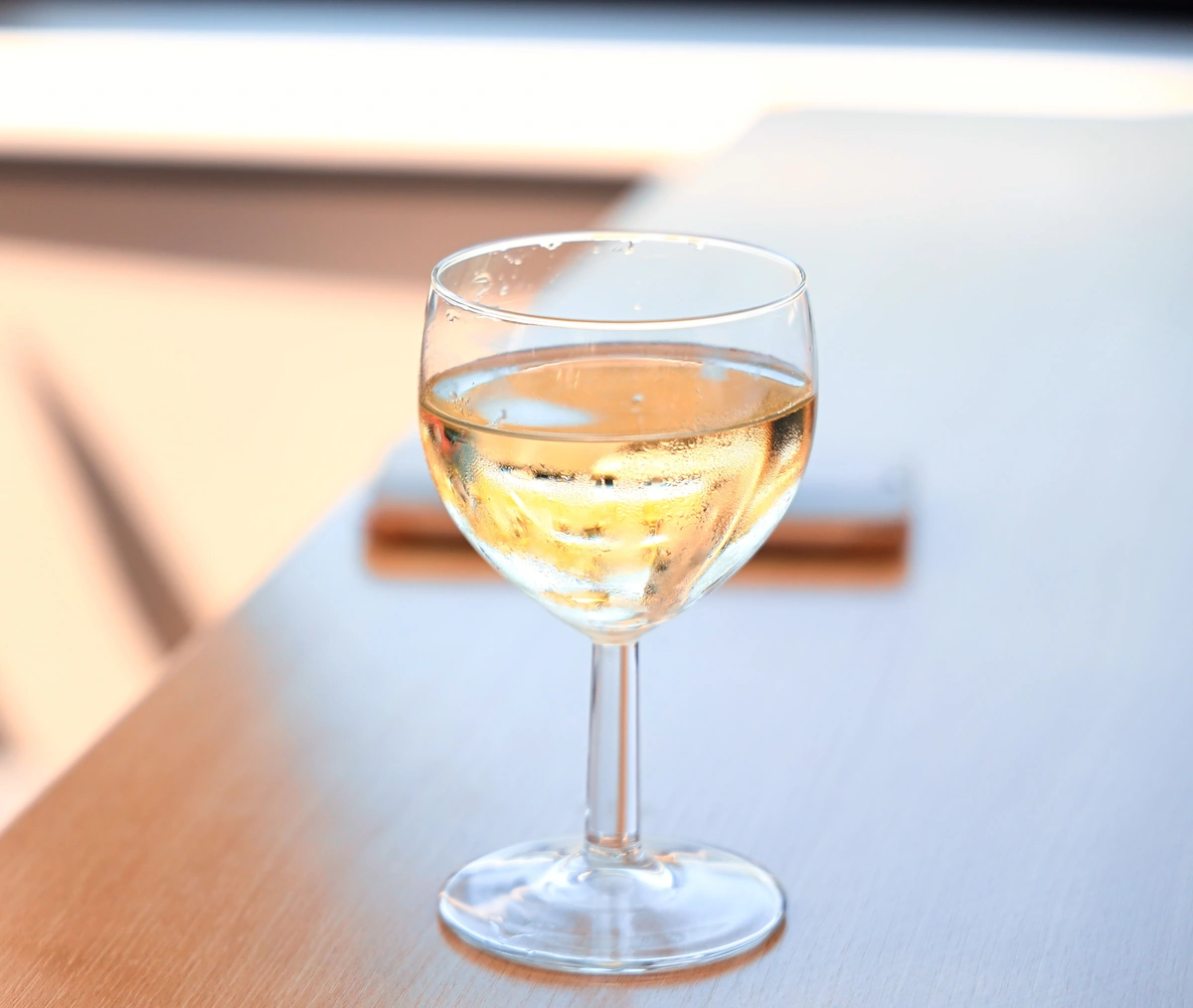 Taça preenchida com vinho branco claro sobre mesa de madeira clara, iluminada por luz natural.