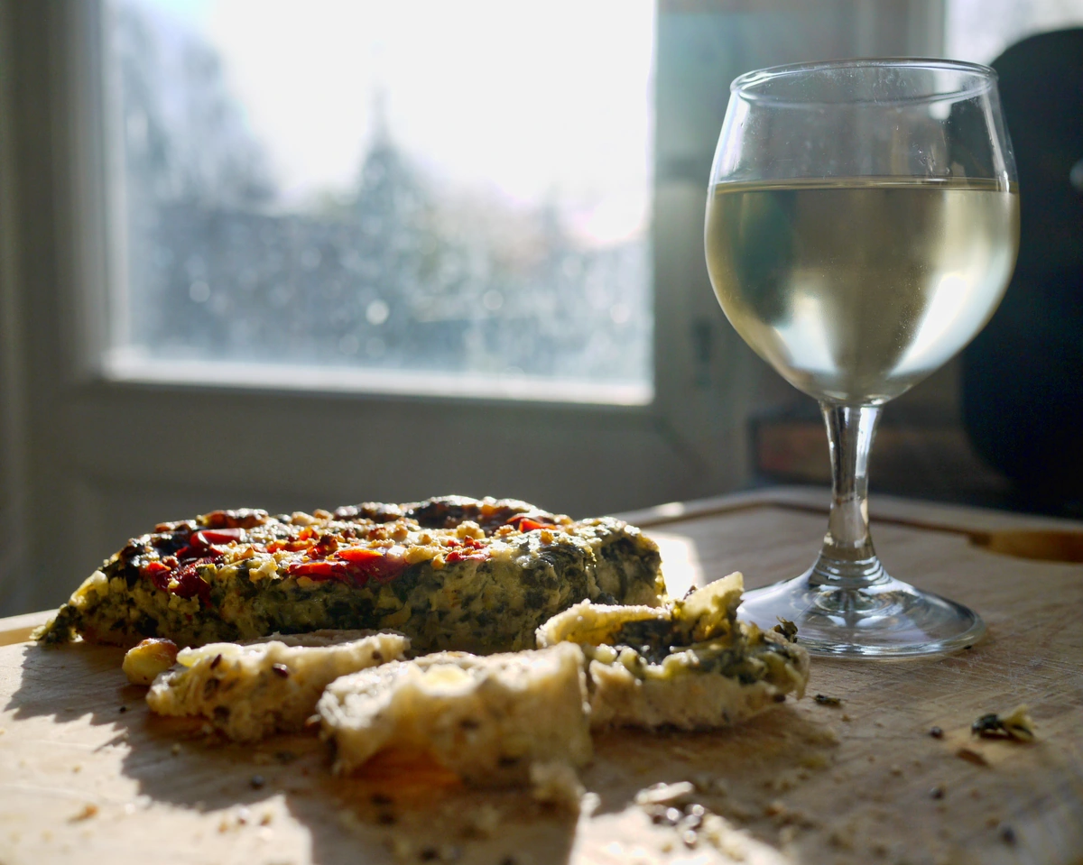 Quiche e pedaços de pão sobre uma tábua ao lado de uma taça preenchida com vinho branco, iluminados pela janela.