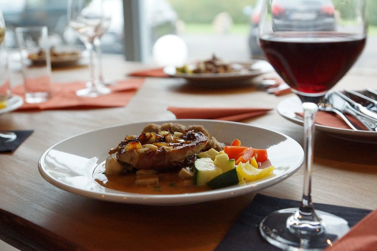 Carne ao molho com legumes coloridos em prato branco, acompanhada de taça preenchida com vinho tinto em mesa de restaurante.