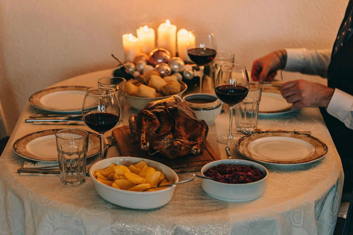 Ave assada no centro de mesa com batatas, molhos e três taças preenchidas de vinho tinto, em jantar iluminado por velas.