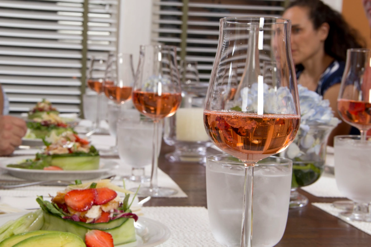 Taça preenchida de vinho rosé diante de uma salada de folhas com pepino e morangos, em uma mesa compartilhada.