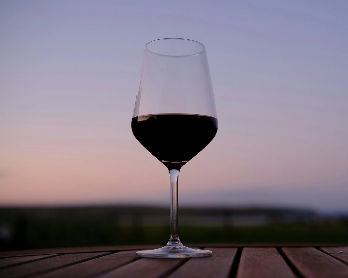Taça preenchida de vinho tinto escuro sobre mesa de madeira, com céu de entardecer ao fundo.