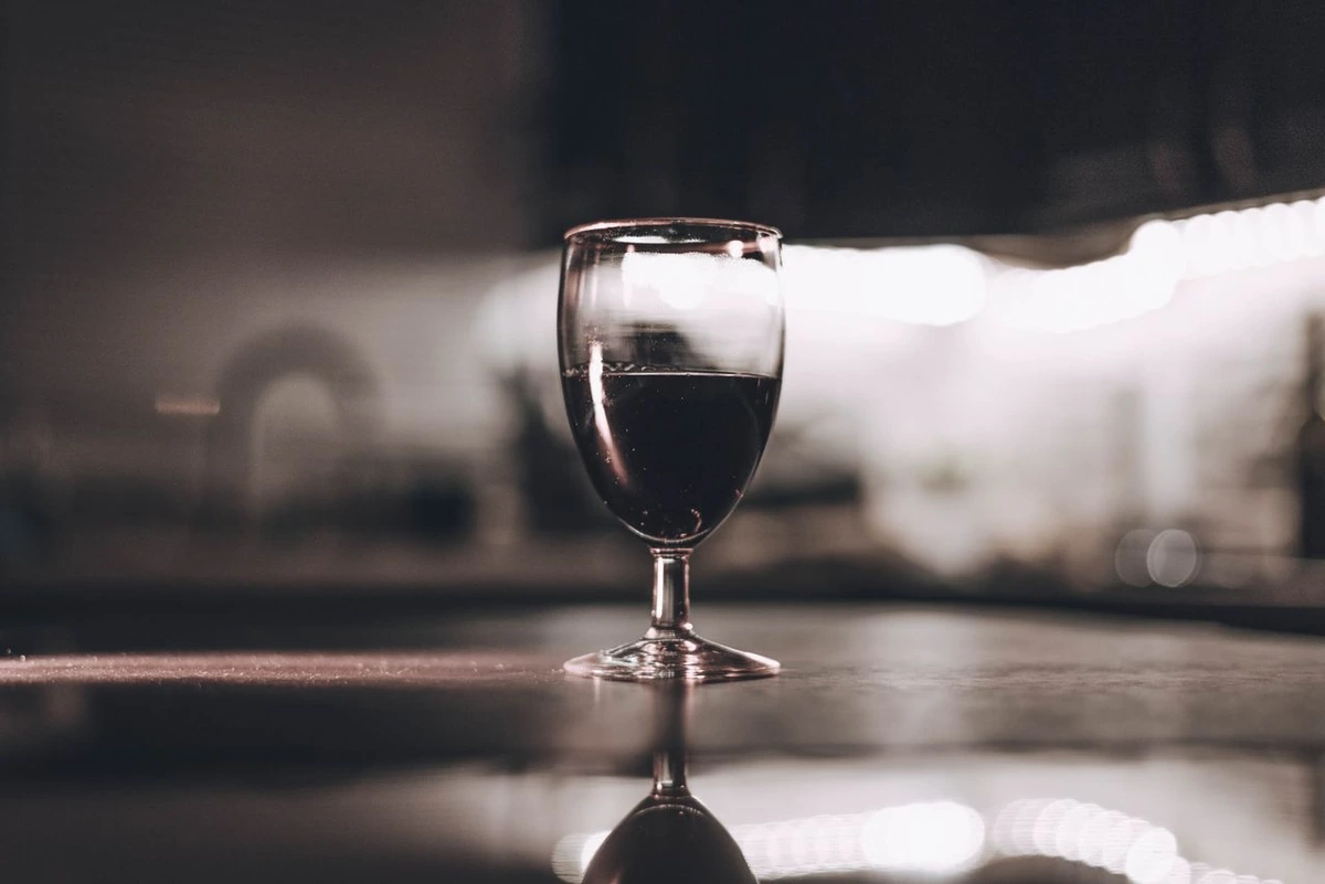 Taça com vinho tinto escuro sobre mesa, com reflexo da taça na superfície e fundo desfocado.