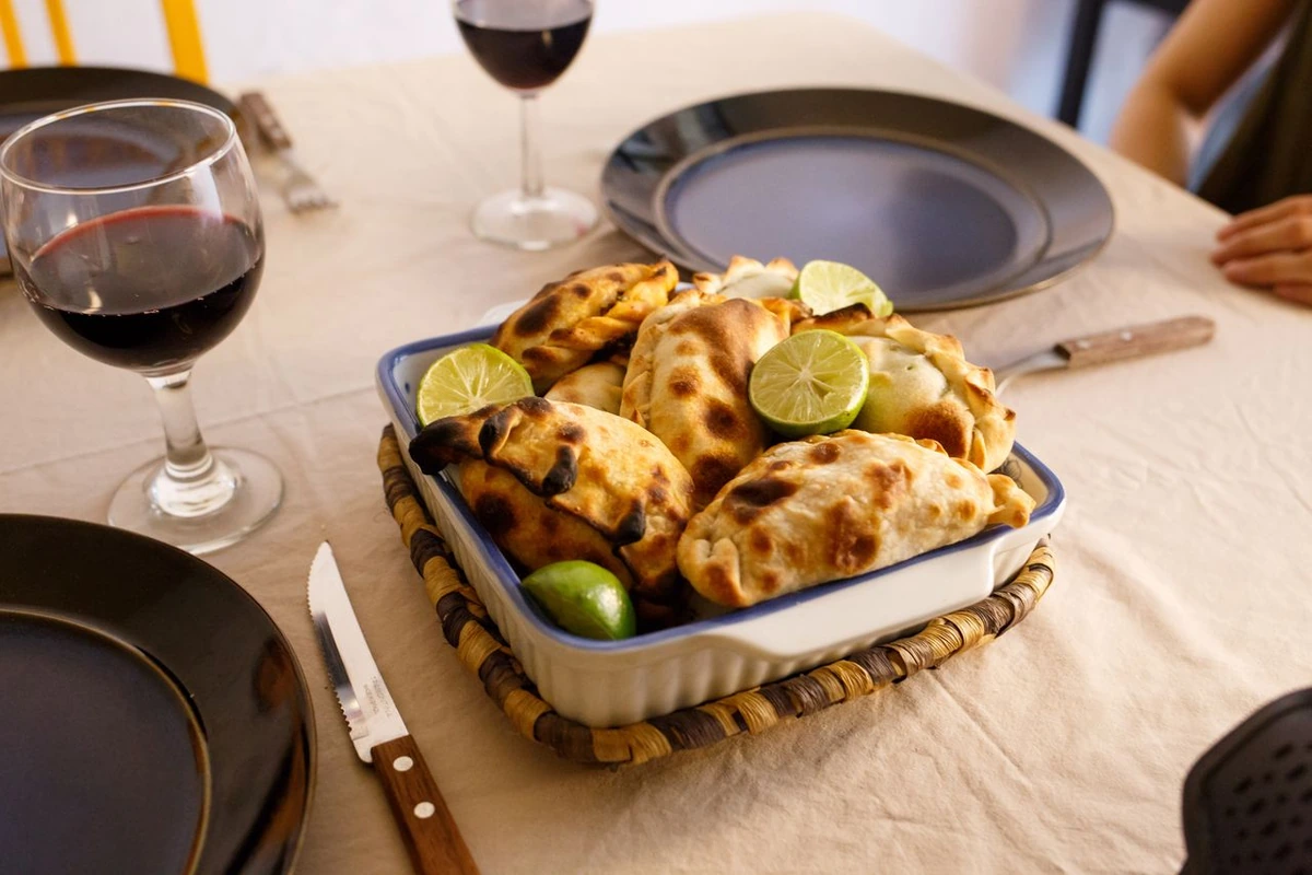 Empanadas douradas em uma travessa com cítricos cortados, acompanhadas de taças preenchidas com vinho tinto.