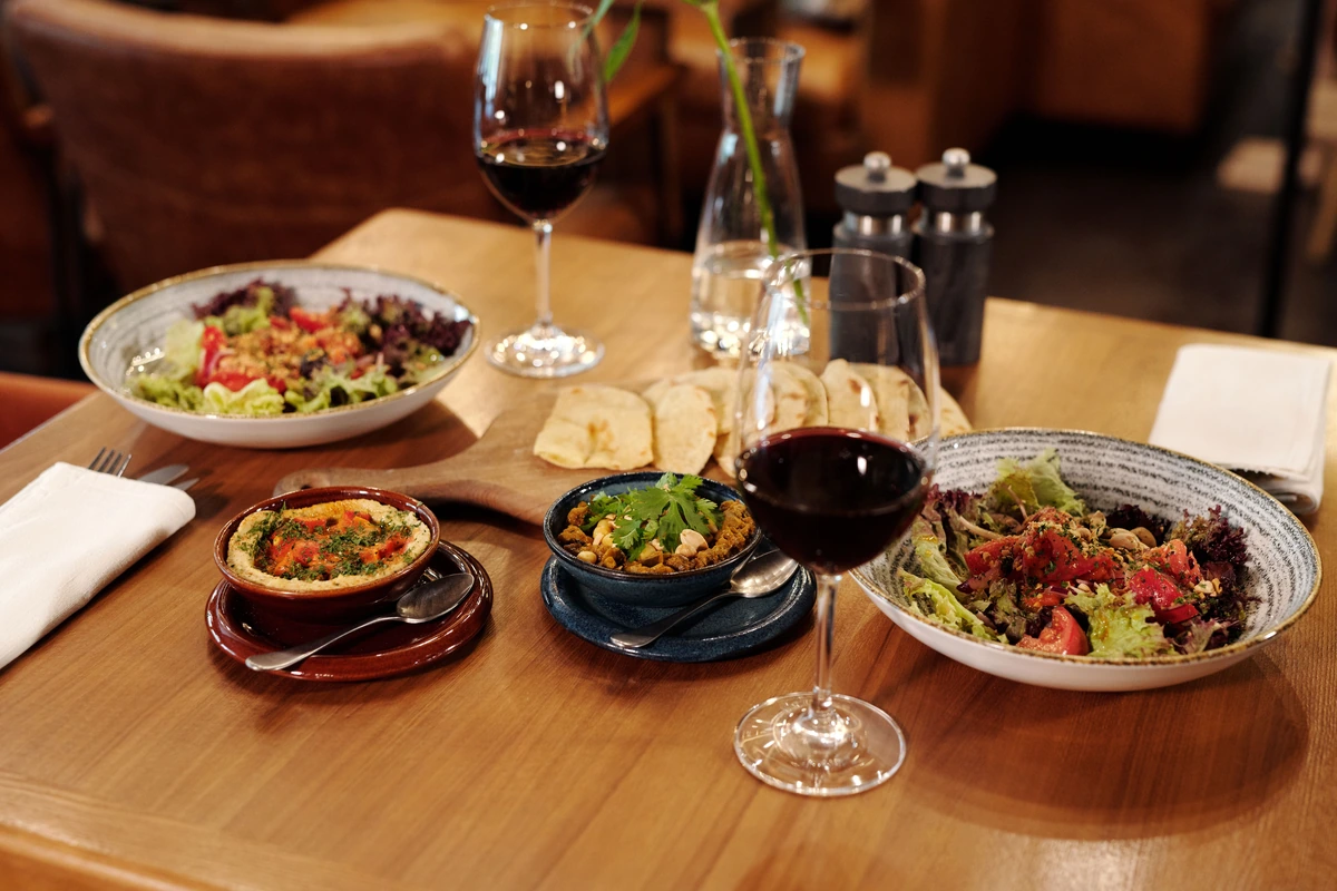 Mesa de madeira com duas taças de vinho tinto, pães, salada e acompanhamentos.