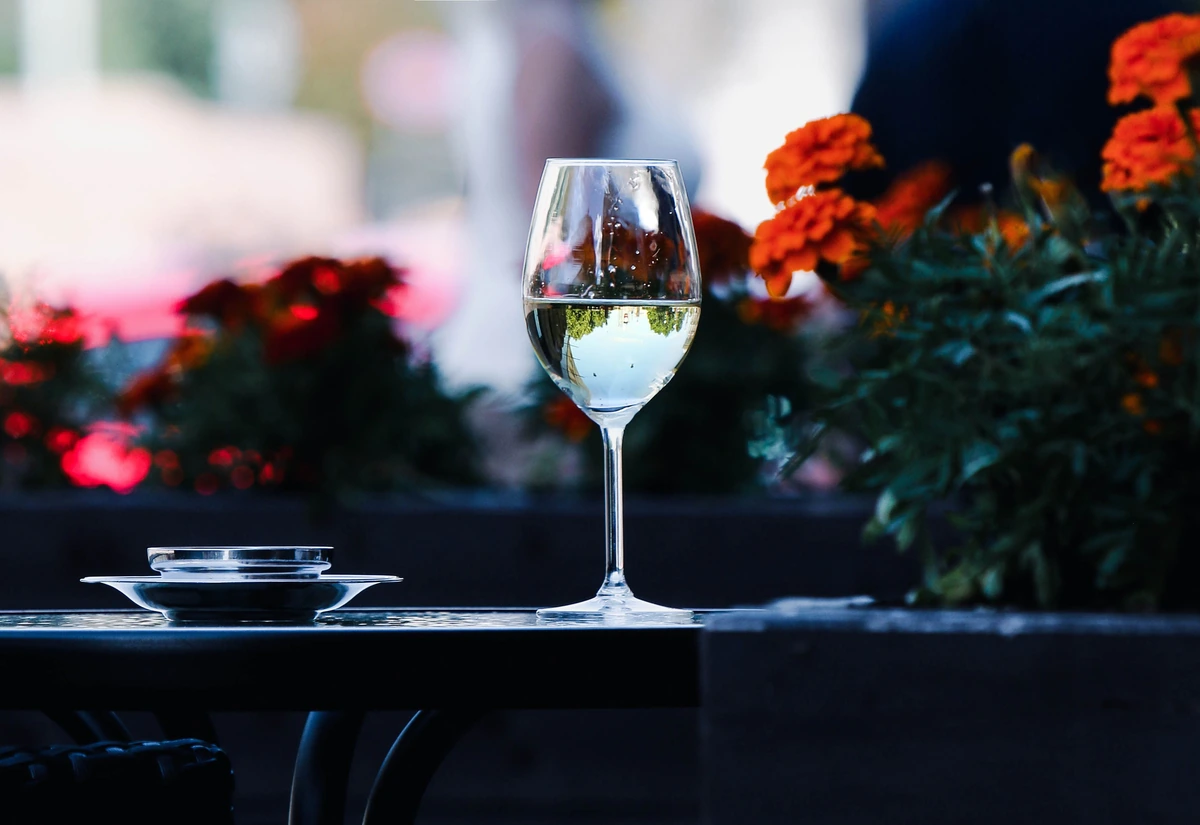 Taça de vinho branco sobre mesa externa, com flores ao fundo.