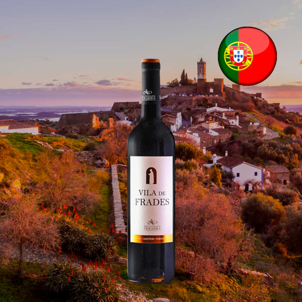 Garrafa de Vila de Frades Tinto, com cápsula preta e rótulo claro, diante de uma vila murada sobre uma colina ao entardecer, com bandeira de Portugal.