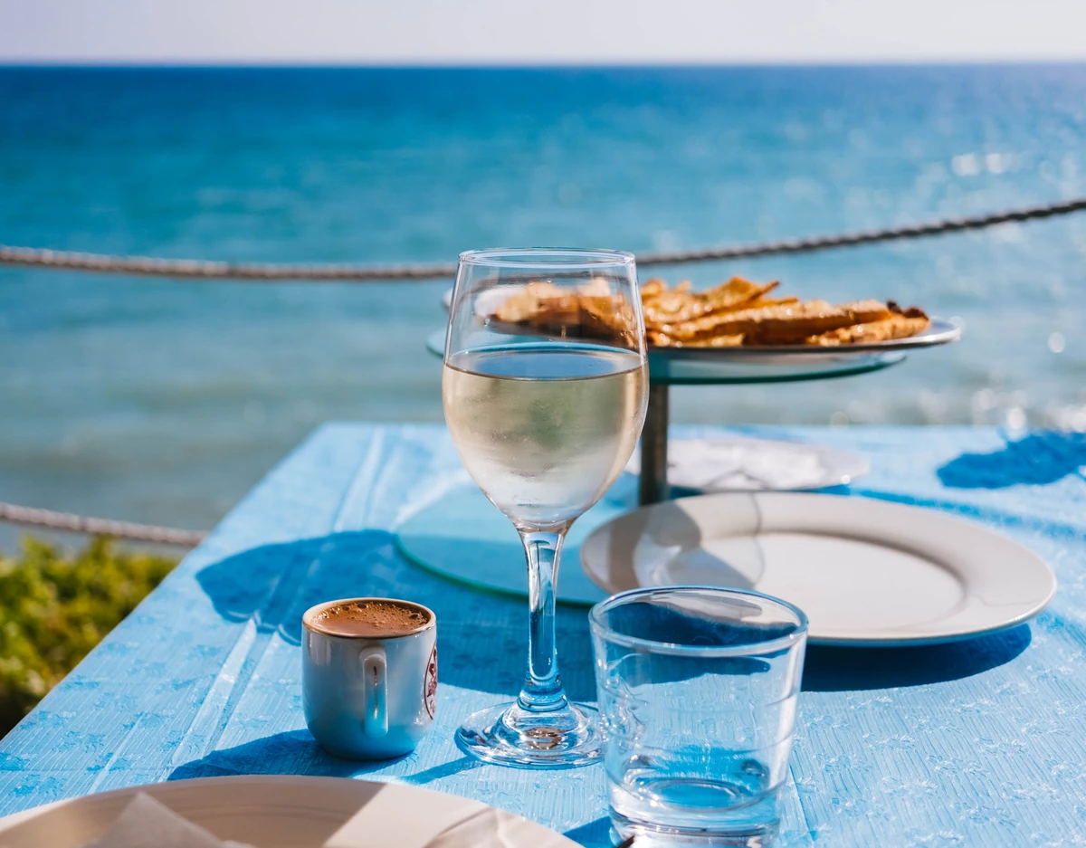 Taça de vinho branco em primeiro plano sobre mesa azul à beira-mar.
