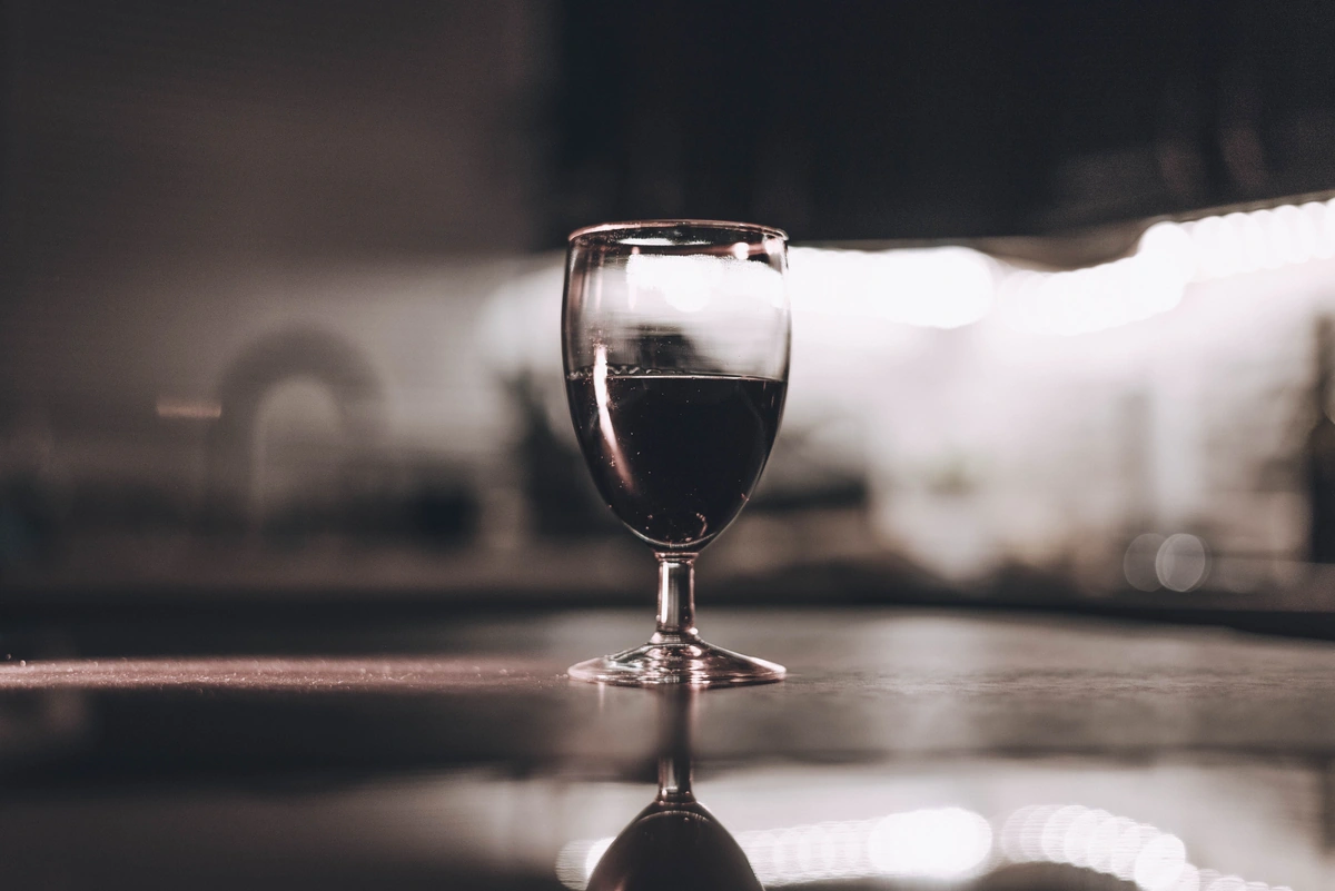 Taça preenchida de vinho tinto sobre mesa escura, com fundo sóbrio desfocado e luz lateral.